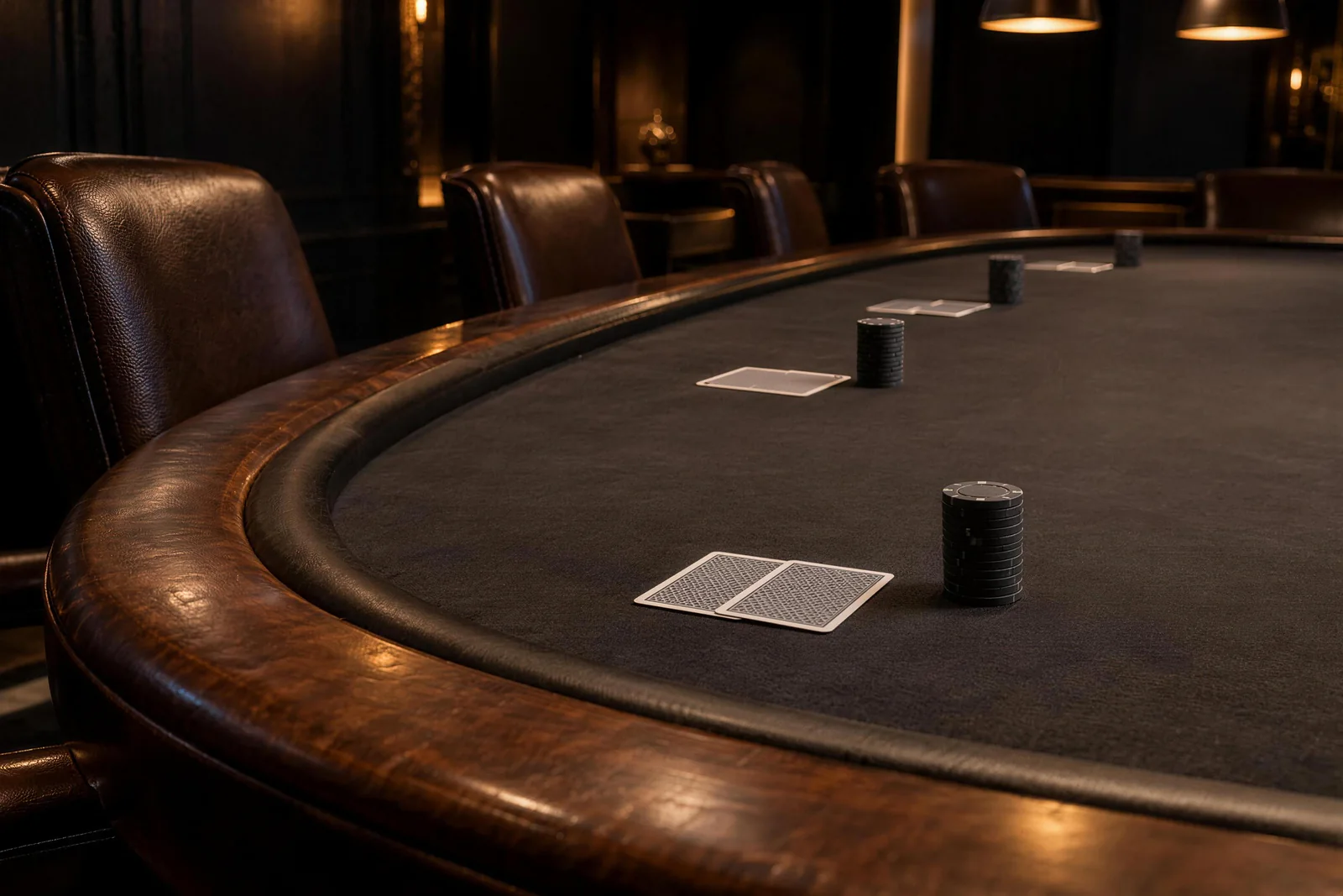 Dark stacks and cards along the felt Paired face-down cards and dark chip stacks line the felt beside empty leather chairs at a poker table
