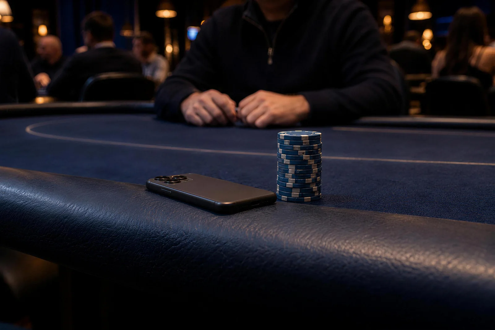 A blue stack beside the phone Near a seated player's hands, a blue chip stack stands beside a face-down phone on the poker rail