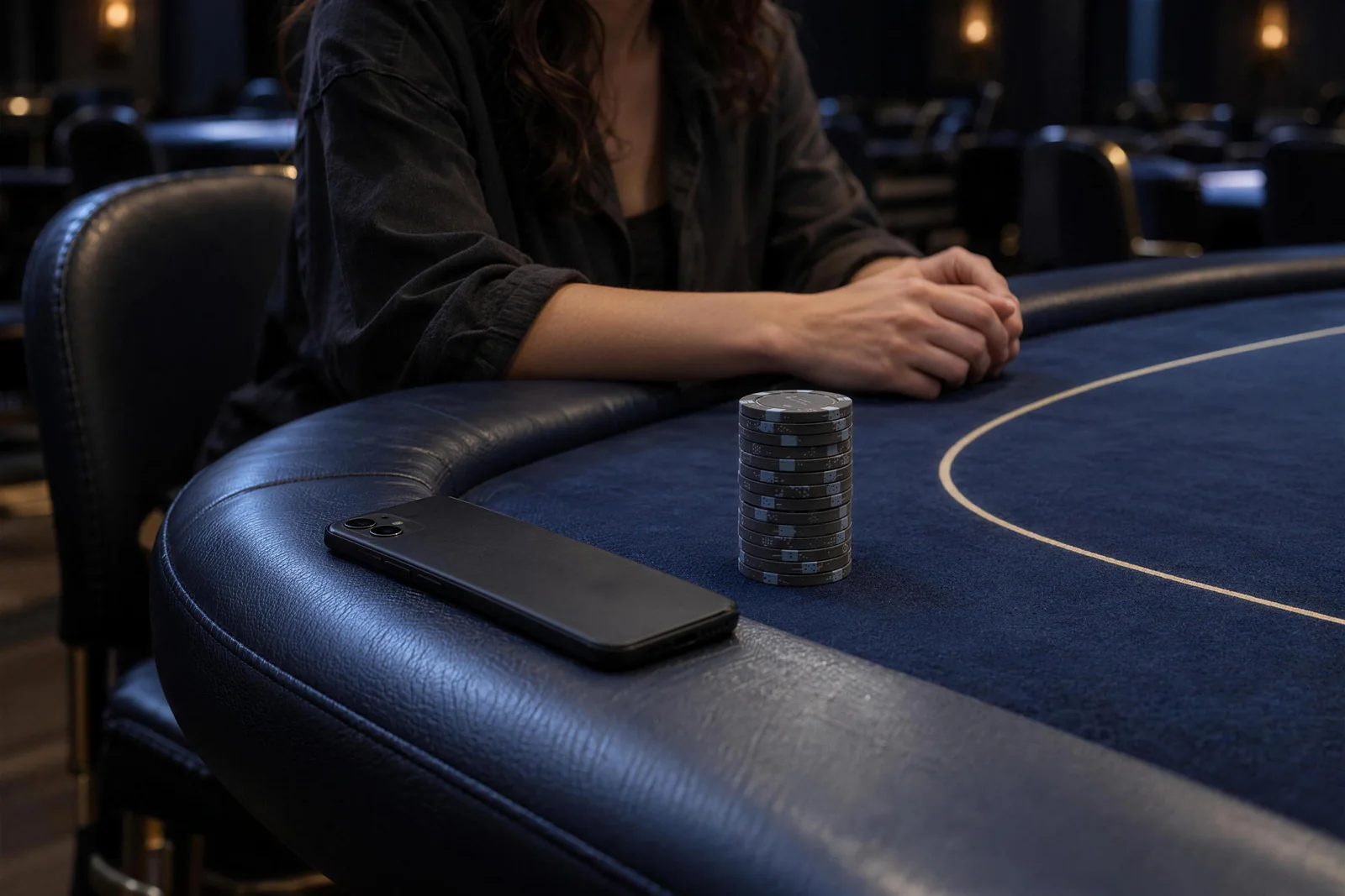 Along a curved poker rail, a face-down phone lies beside a dark chip stack and a seated player's arms